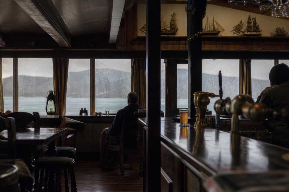 Interior of a small island pub with dark timber bar, brass fittings, model ships on a shelf and rain streaking the window behind