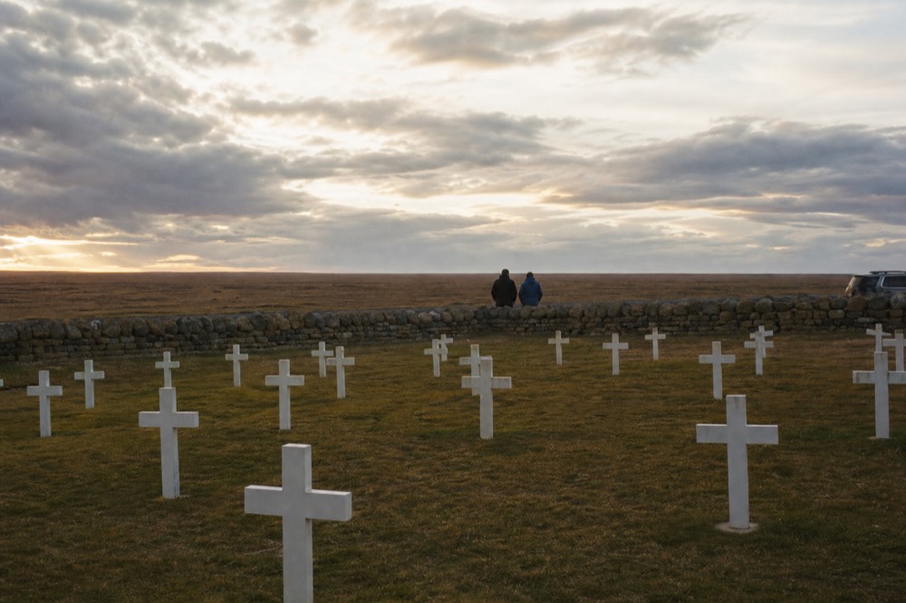 Rows of plain white crosses on close-cropped grass inside a low stone wall, open moorland stretching flat to the horizon behind