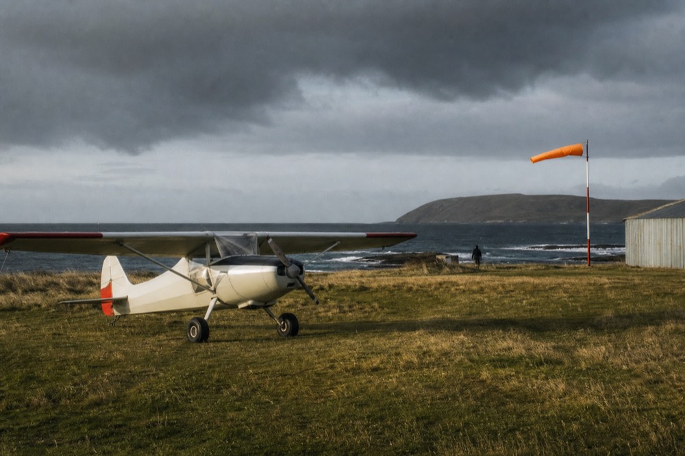 Small propeller aircraft parked on a grass airstrip beside a wind sock, low hills and a corrugated iron shed in the background