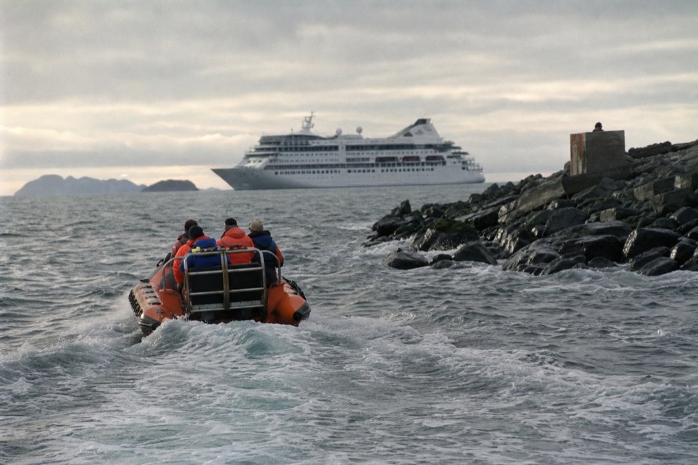 Orange tender boat crossing choppy grey water toward a stone jetty, a large white cruise ship anchored out in the sound behind it