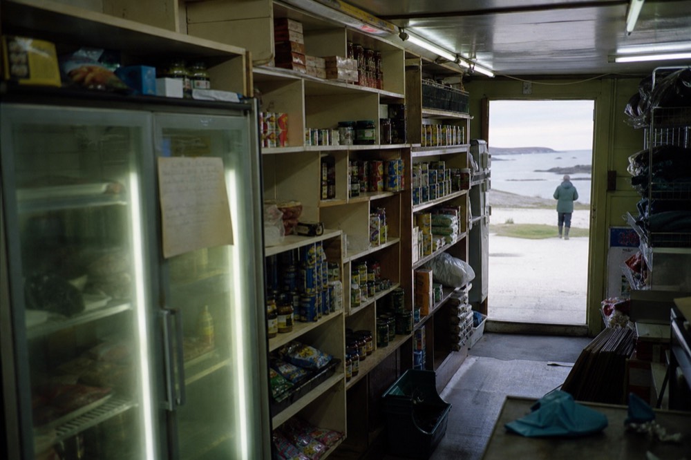 Small general store interior with tightly packed shelves of imported tins and packets under strip lighting, a hand-written notice taped to a chilled cabinet door