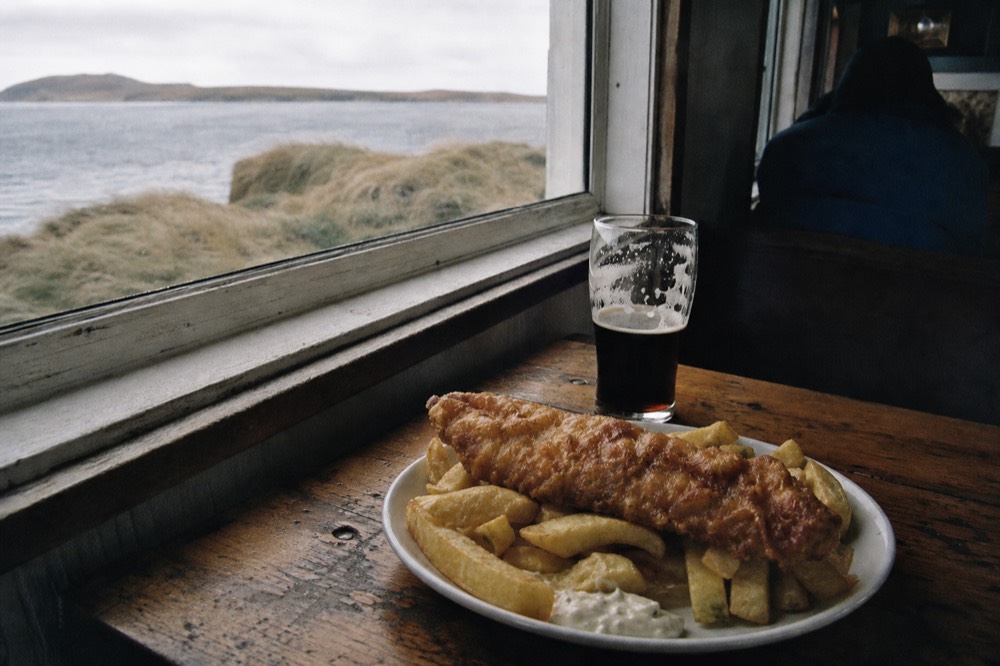 Plate of battered fish and thick-cut chips on a scarred wooden pub table beside a dark half-finished pint, low afternoon light through a small salt-hazed window