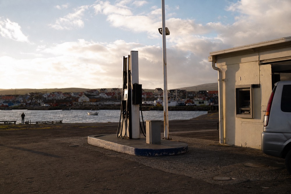 Fuel station forecourt at the edge of a small town on a bright windy afternoon, single pump island and a low utilitarian building with a cash machine set into the wall