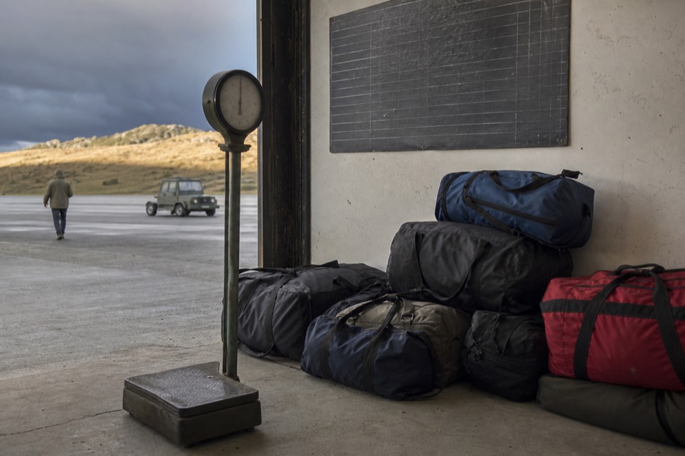 Luggage scales and a stack of soft holdalls on the concrete floor of a tiny airstrip building, a hand-chalked weight board on the wall behind