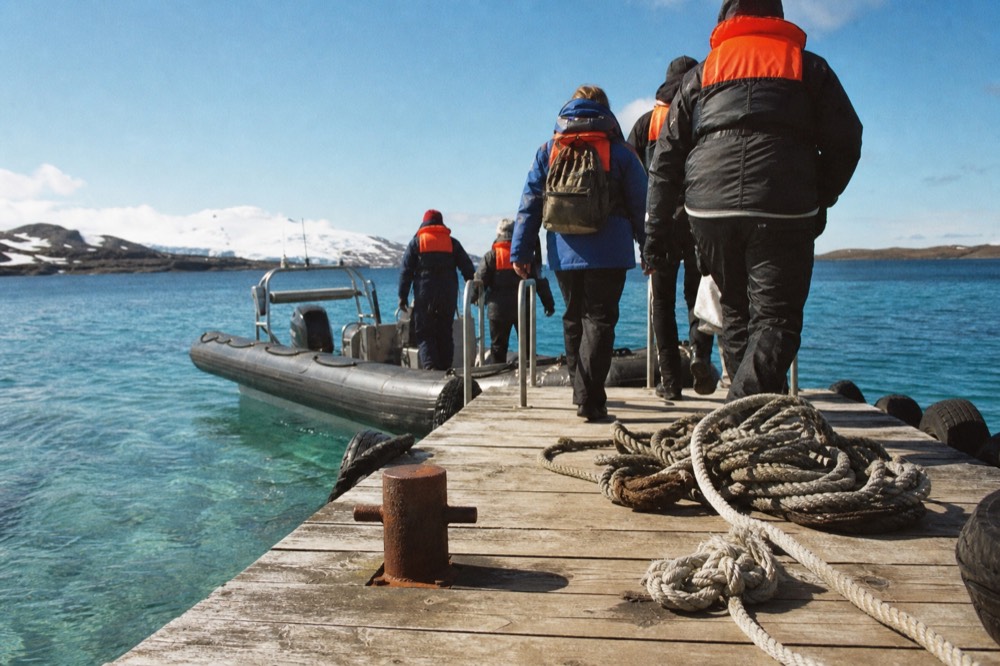 Passengers in life jackets stepping from a tender onto a floating pontoon in bright low sunshine, coils of rope and a rusted bollard on the weathered jetty planking