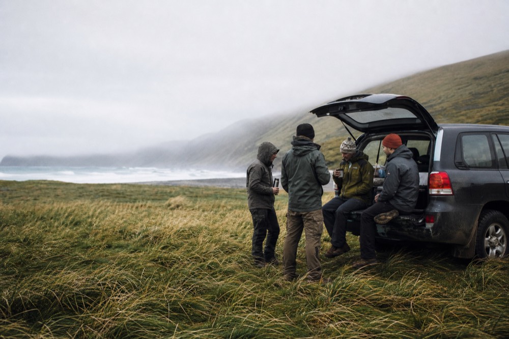 Four travelers in layered outdoor clothing sharing hot drinks from flasks on the tailgate of a parked 4x4, wind flattening the grass around them