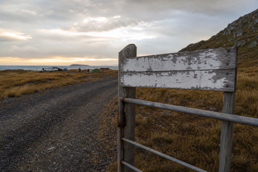 Hand-painted wooden sign on a gatepost beside a gravel track, chipped white lettering weathered by salt wind with open camp stretching behind it