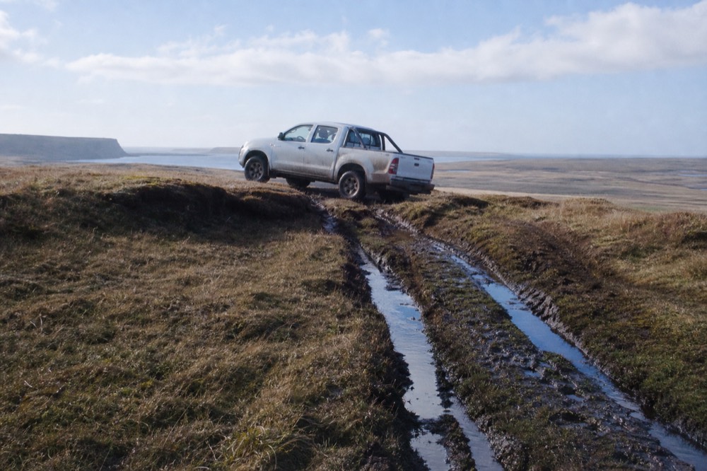 A four-wheel-drive pickup nosing down a peat bank onto open grass, deep water-filled wheel ruts behind it, no road anywhere in frame
