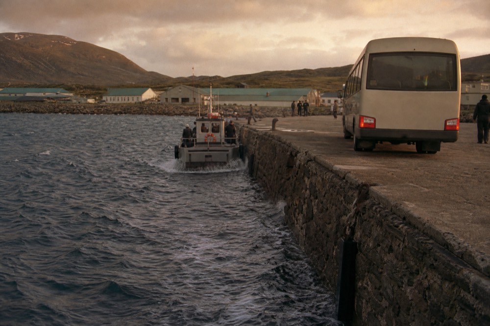 A stone jetty at the edge of a small harbour with a minibus waiting on the quay and a tender coming alongside in choppy water